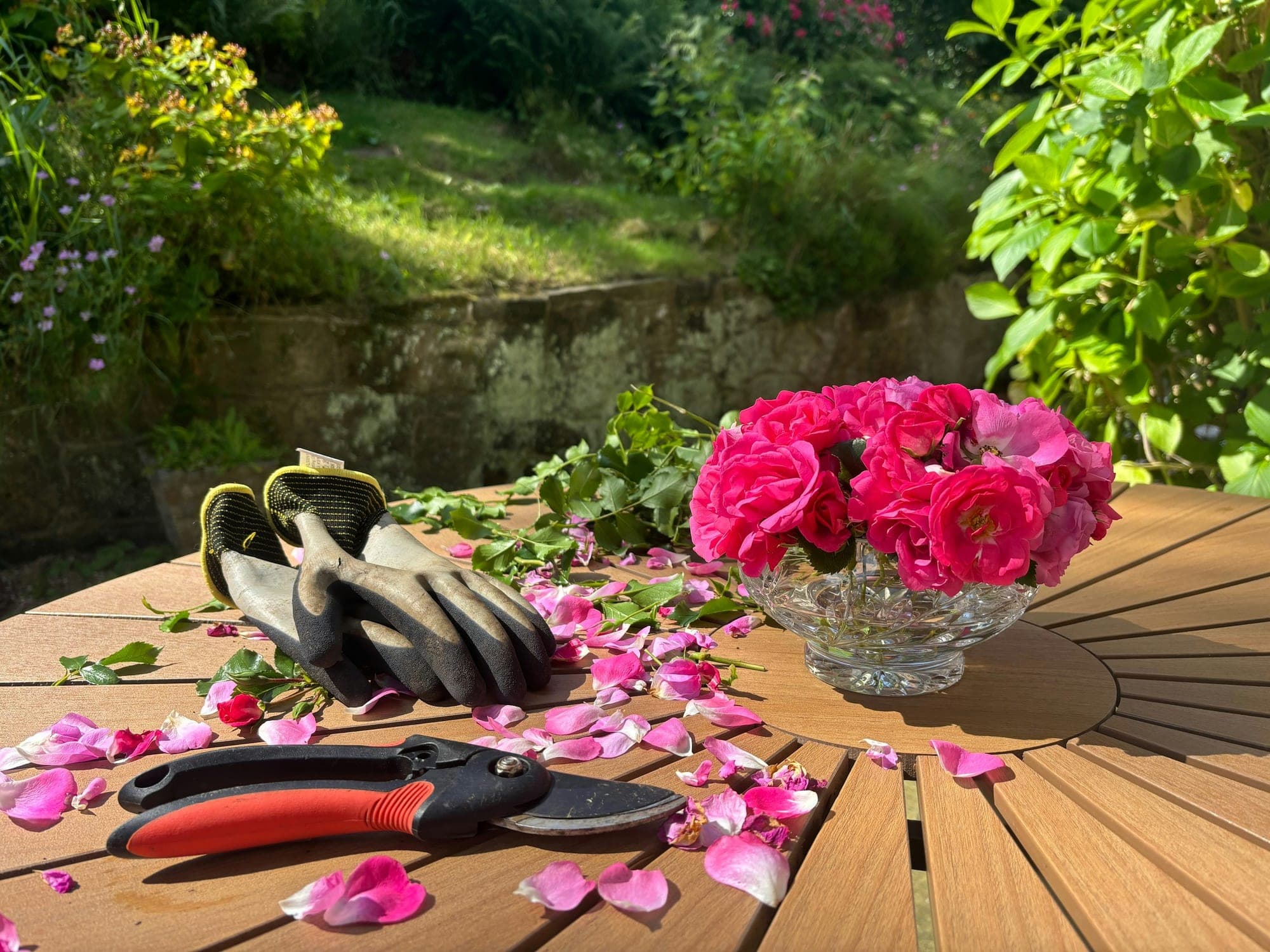 Red-handled secateurs and a pair of gardening gloves on a garden table beside a bowl of pink roses, with a sunny planted bank behind.