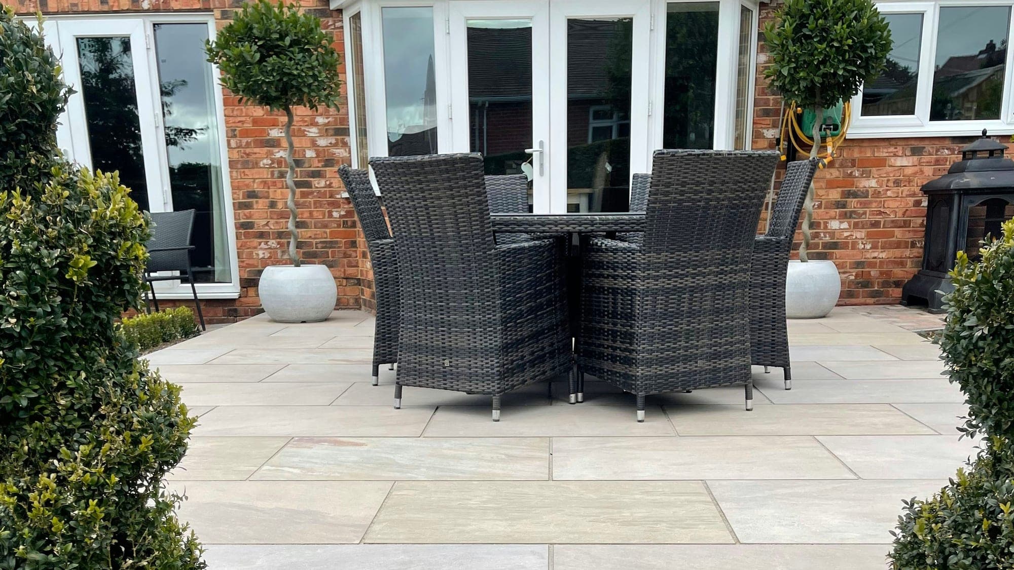 A new pale porcelain-paved patio with a rattan table and chairs outside a red brick house, flanked by clipped topiary shrubs.