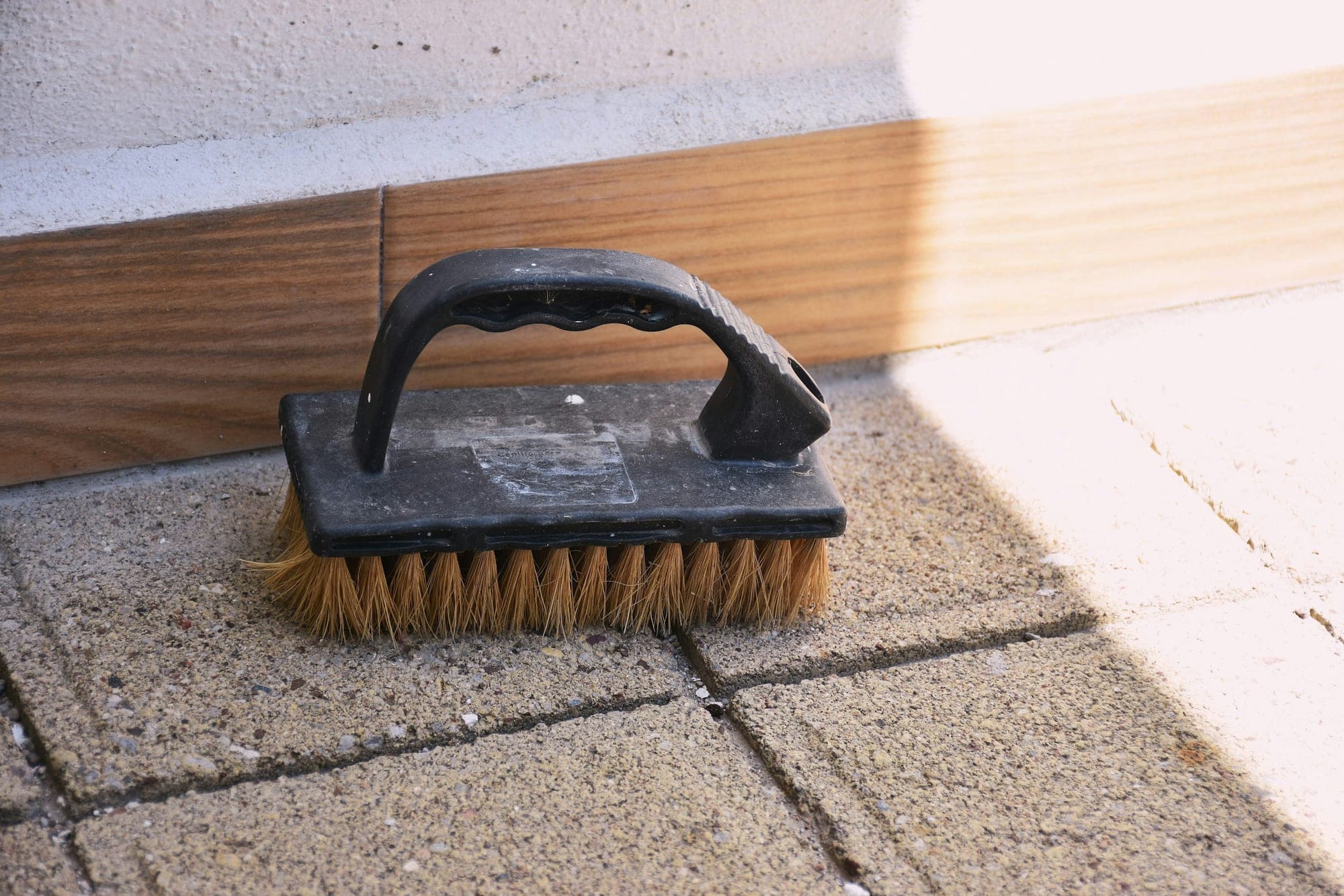 A stiff scrubbing brush resting on paving slabs beside a sunlit wall.