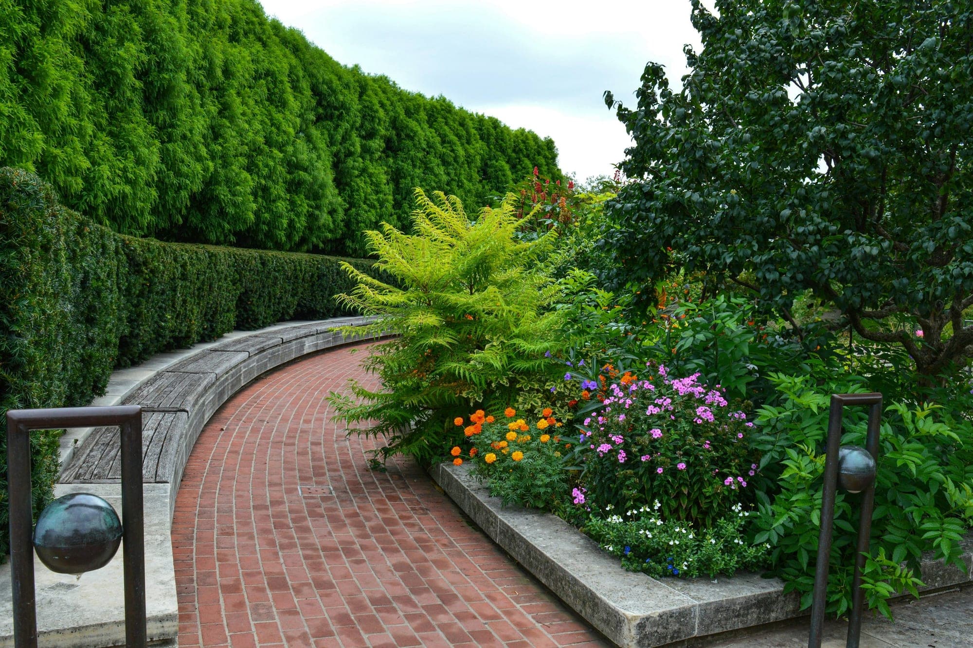 A curving brick-paved path beside a raised stone-edged flower border, with a tall clipped hedge behind.